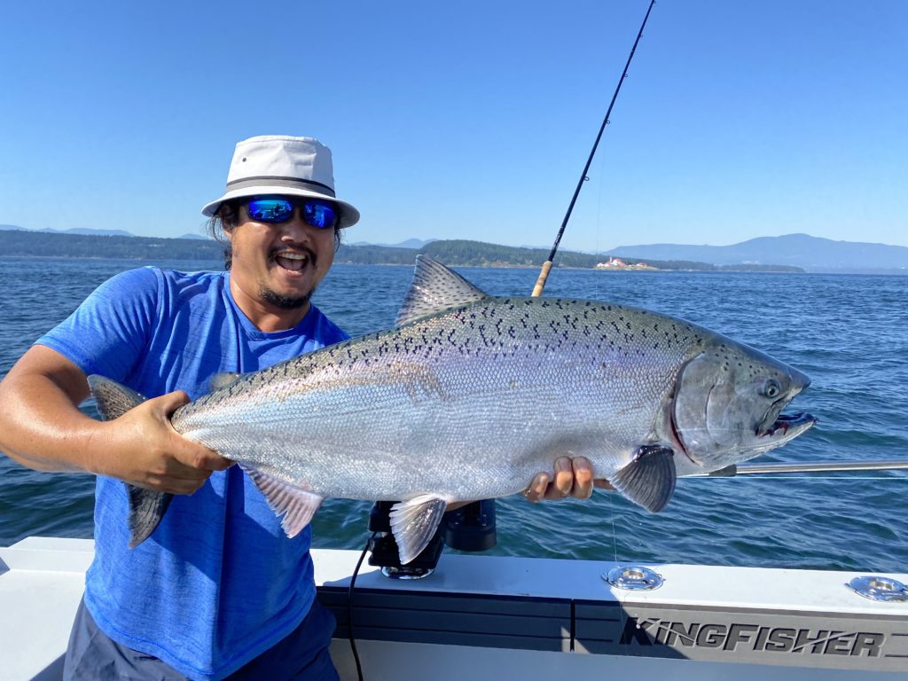 A man in a blue t-shirt and bucket hat holding a gigantic salmon on a sunny day